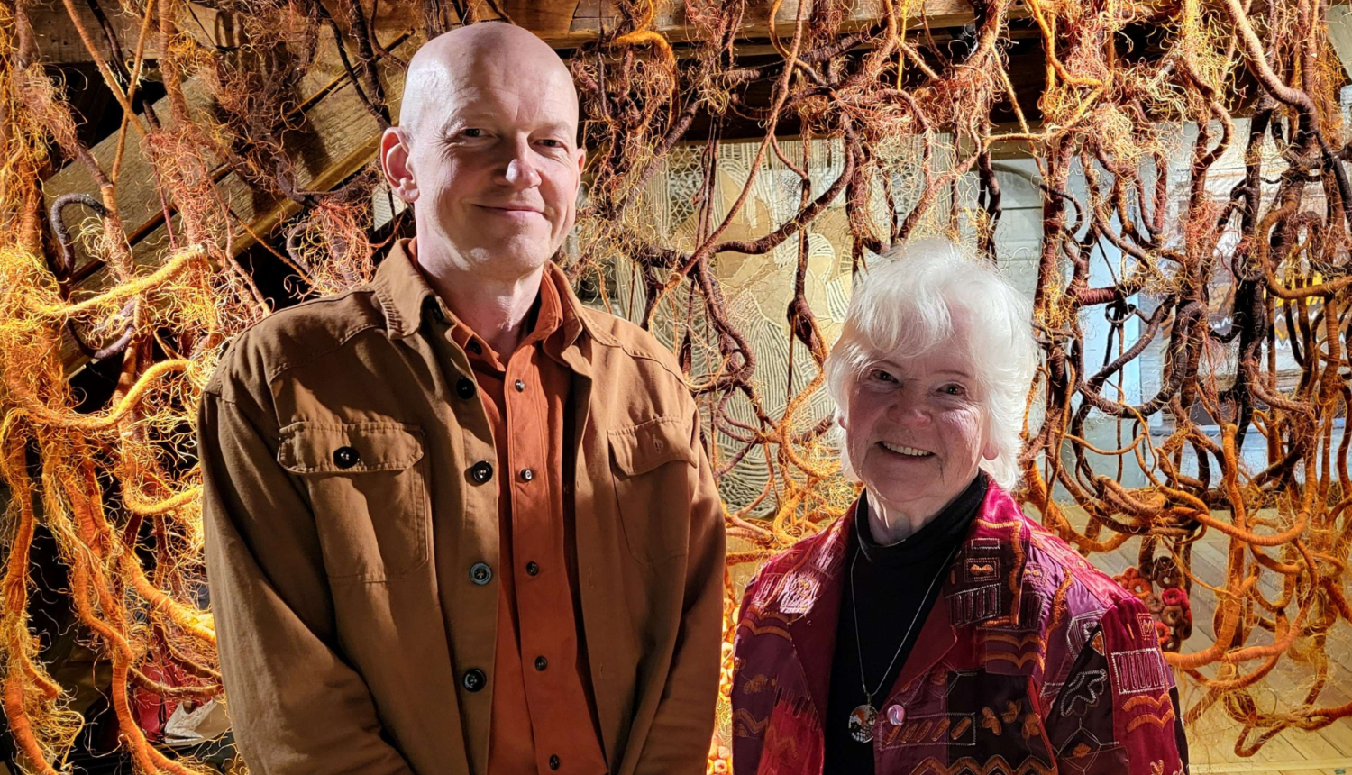 Tekstilmāksliniece Vija Jansone un mākslinieks Kaspars Lielgalvis izstādē “Uzkāpt kalnā”, fonā tekstilmākslas darbs