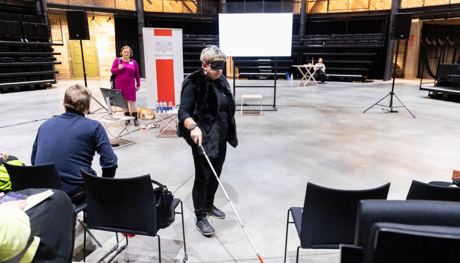Cilvēks ar spieķi auditorijas priekšā demonstrē piekļūstamības izaicinājumus